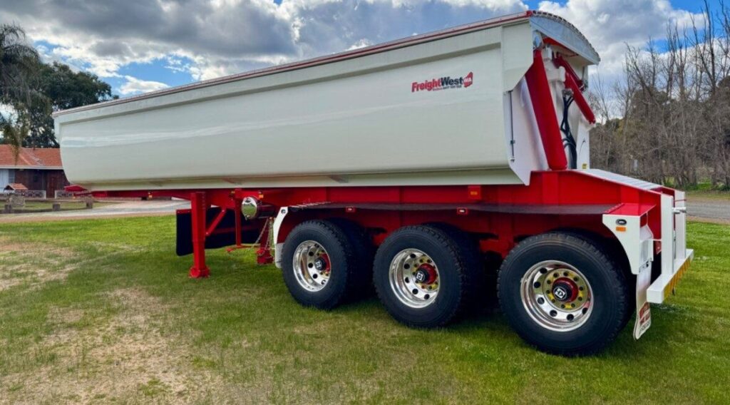 A white and red tri-axle tipper trailer is parked on grass near a road, with trees and a house in the background under a partly cloudy sky. The trailer has the "Freight West" logo on the side. ETA Trailers