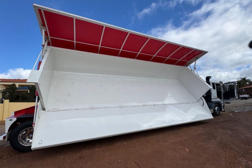 A large white lorry with an open, red-roofed cargo bed is parked on gravel, showing its sideways tilting mechanism for unloading. The sky is partly cloudy. ETA Trailers