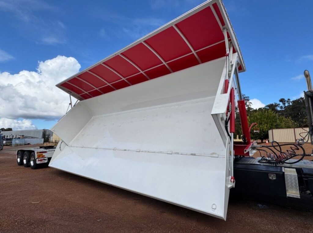 A large white tipper lorry with a red interior roof is raised and tilted, showing its empty cargo area. The trailer is attached to a black lorry on a dirt lot under a partly cloudy sky. ETA Trailers