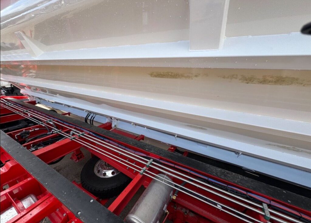 A close-up view of the underside of a white boat on a red trailer, showing some yellowish stains along the hull and various black and red cables running parallel beneath the boat. ETA Trailers