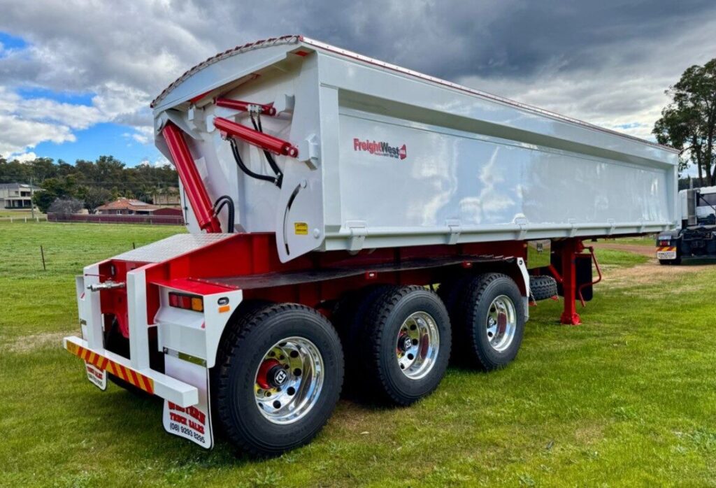 A white and red tri-axle tipper trailer is parked on grass, with polished chrome wheels and hydraulic lift, under a partly cloudy sky. Trees and houses are visible in the background. ETA Trailers