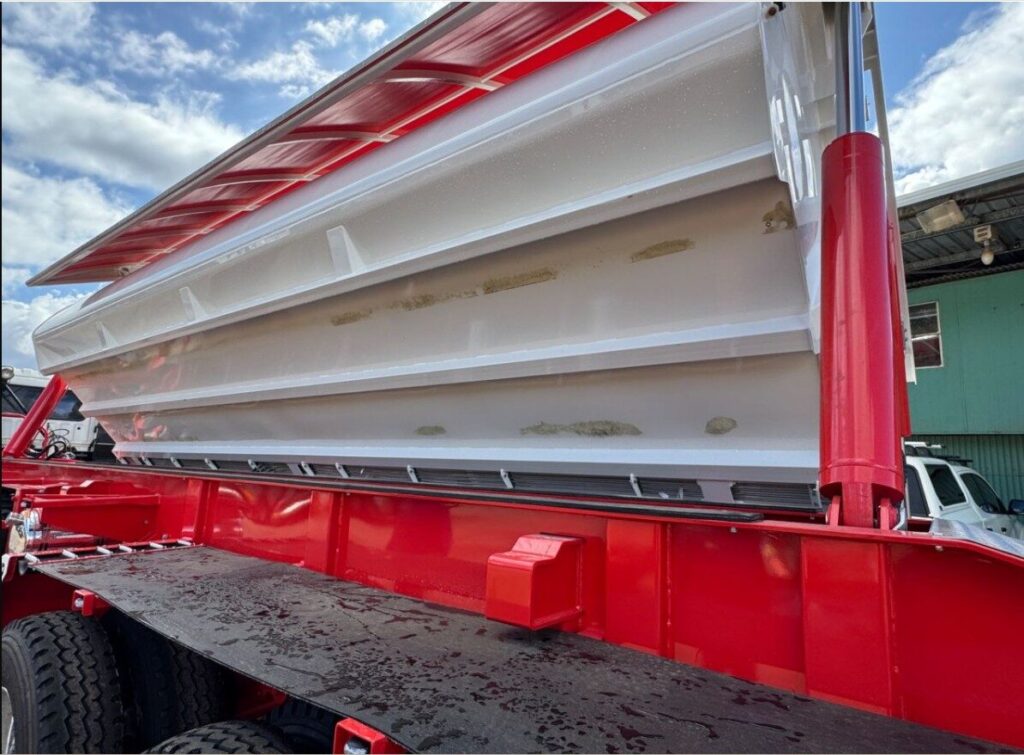 A close-up view of a red tipper lorry with its white bed raised, exposing the underside. The sky is partly cloudy, and part of a building and another vehicle are visible in the background. ETA Trailers